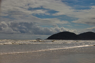 quiet beach, with small waves breaking in a sparkling sea, with hills in the background, under a dramatic sky covered by large white clouds.