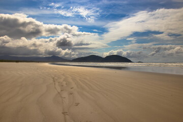footprints heading towards the horizon, imprinted on the golden sands of the sparkling water beach, with mountains in the background under a blue sky with large white clouds.