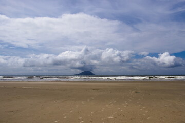 footprints towards the sea with small waves marked from the humid sand of the beach, on the horizon a small island framed by a blue sky with white cumulus clouds.