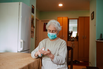Portrait of a cute senior grandmother who pours an antibacterial hand sanitizer into her hand while sitting at a table in the house. Hand hygiene during isolation of COVID-19 coronavirus