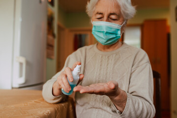 A Caucasian senior woman with a face mask who pours an antibacterial hand sanitizer into her hand while sitting at a table in the house. Hygiene during the COVID-19 coronavirus pandemic