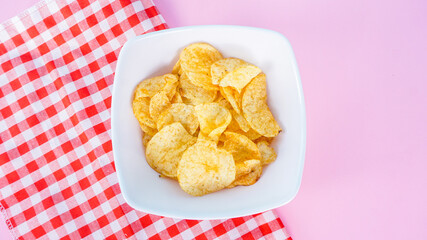 Naklejka premium Potato chips in bowl on napkin and pink background, top view. Salty crisps scattered on a table