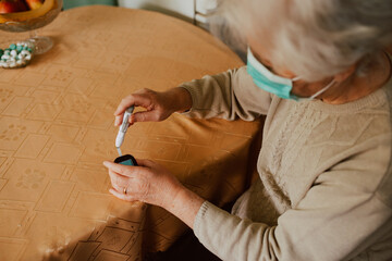 Top view of a senior grandmother with a face mask holding a blood sugar meter in her hand while...