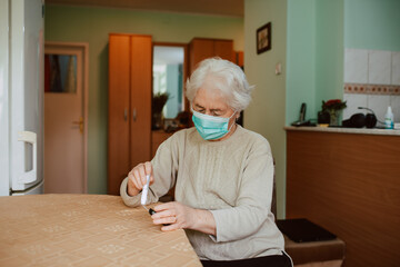 A senior grandmother with a face mask holding a blood sugar meter in her hand while sitting at a...