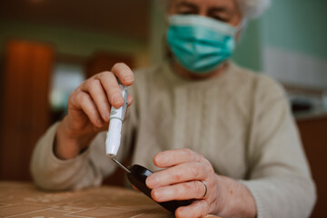 A retired senior woman with a face mask holding a blood sugar meter in her hand. Health prevention during the COVID-19 coronavirus pandemic