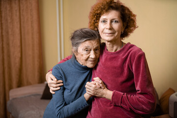Elderly old cute woman with Alzheimer's very happy and smiling when eldest daughter hugs and takes care of her, at home on sofa. Theme aging and parenting, family relationships and social care oldest