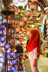 Young pretty tourist Muslim woman with red scarf looking on Traditional Turkish lamps in souvenir shop. Woman in hijab shopping in the Grand Bazar. Shopping tour.