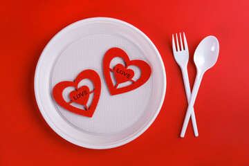Top view of plastic white disposable fork, spoon, plate and red hearts isolated on red background, valentine's day for poor people concept