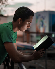 Hispanic man reading a black book at sunset while his dark face lights up