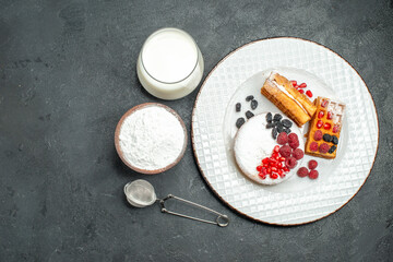 top view tart and waffles with berries on white oval plate a bowl of powdered sugar a sieve and a glass of milk on dark background with copy space