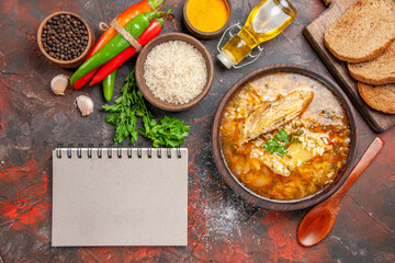 top view chicken and rice soup in a bowl wooden spoon hot pepper bottle of oil parsley bowl of rice slices of bread on chopping board different spices a notebook on dark red background