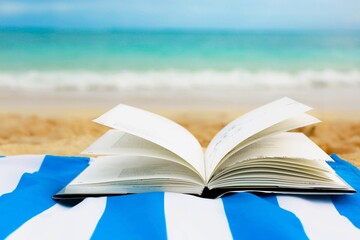 A opened book on the beach sand in front of a blue ocean. Literature and relaxation.