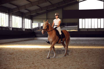 Young woman in equestrian suit riding horse indoors. Beautiful pet