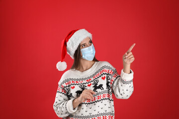 Fototapeta na wymiar Pretty woman in Santa hat and medical mask pointing on red background