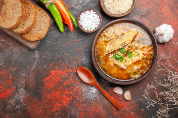 top view chicken and rice soup in a bowl wooden spoon hot peppers bowls with sea salt and rice garlic slices of bread on chopping board on dark red background with copy space