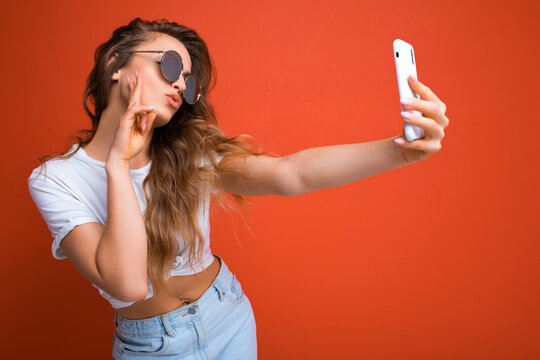 sexy beautiful young blonde woman holding mobile phone taking selfie photo using smartphone camera wearing sunglasses everyday stylish outfit isolated over colorful wall background looking at device