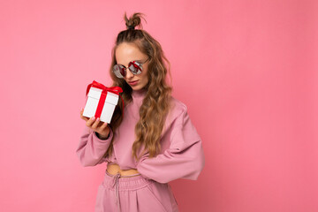 Attractive happy adult blonde curly woman isolated over pink background wall wearing pink sport clothes and sunglasses holding gift box looking at present
