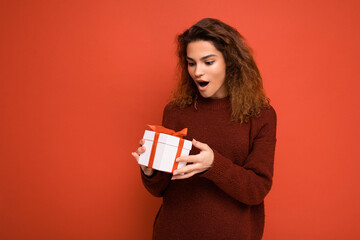 Attractive positive surprised young brunette curly woman isolated over red background wall wearing white sweater holding gift box and unboxing present looking at box with red ribbon