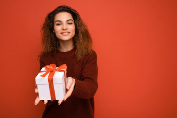 Beautiful happy young brunette woman isolated over colourful background wall wearing stylish casual clothes holding gift box and looking at camera