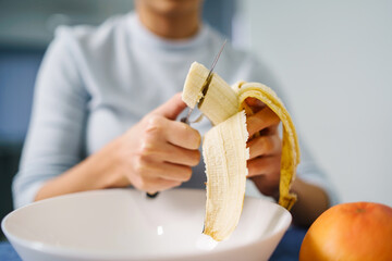 Caucasian woman sitting by the table at home cutting banana - Adult girl female preparing fruit...