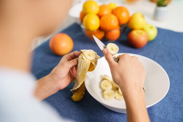 Close up on hands of unknown caucasian woman cutting banana by the table at home - back view top...
