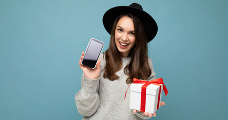 Shot of pretty smiling positive young brunette woman isolated over blue background wall wearing stylish black hat and grey sweater holding gift box showing smartphone screen display for mockup and