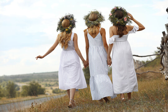 Young women wearing wreaths made of beautiful flowers outdoors on sunny day, back view