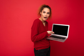 Close-up portrait of beautiful amazed surprised astonished dark blond woman holding laptop computer looking at camera showing at netbook keyboard wearing red sweater isolated over red wall background