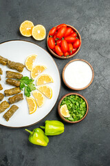 Obraz na płótnie Canvas top distant view stuffed grape leaves parsley leaves and lemon half slices on white oval plate bowls with cherry tomatoes natural yogurt parsley and green peppers on dark background