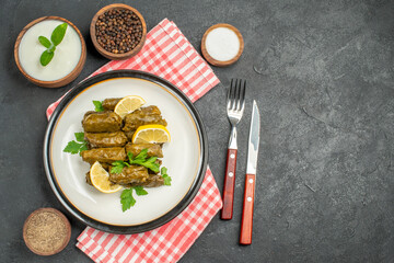 top view stuffed grape leaves on white plate bowls of natural yogurt black pepper salt pepper knife and fork napkin on dark background