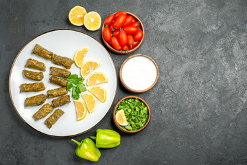 top view stuffed grape leaves parsley leaves and lemon half slices on white oval plate bowls with cherry tomatoes natural yogurt parsley and green peppers on dark background