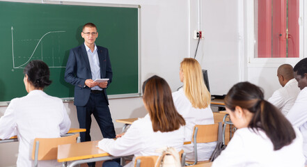 Portrait of teacher standing near chalkboard, giving lesson to group of students as part of adult education course
