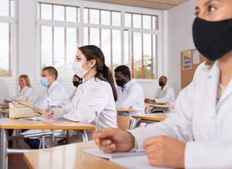 Interested female medical student wearing protective face mask making notes during group lecture in classroom. Necessary precautions in public place during coronavirus pandemic