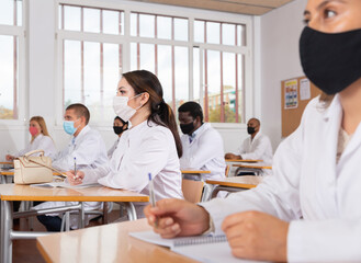 Fototapeta premium Interested female medical student wearing protective face mask making notes during group lecture in classroom. Necessary precautions in public place during coronavirus pandemic