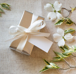 A gift box wrapped in festive silver with white bow and blank name tag with white spring flowers.  It's square with top down view.