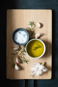 Bowls of salt flakes and olive oil with rosemary and garlic cloves on a wooden cutting board.