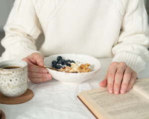 Reading book and eating healthy holiday winter breakfast with granola muesli and yogurt in bowl on white table background. Organic morning diet meal with oat