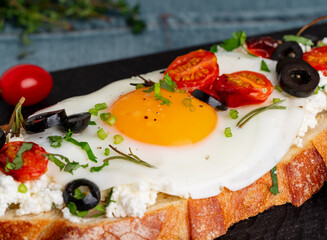Toasted bread toast with fried eggs with yellow yolk and tomatoes, olives, sprinkled with herbs on dark wooden serving board on blue denim napkin, side view, close up, macro