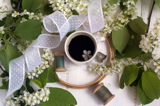 spring flower background: bird cherry, cup of coffee, hoop, spools of thread, nylon white ribbon on wooden background