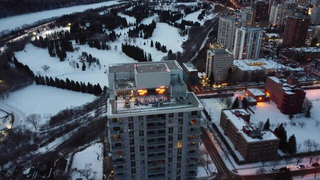 aerial birds eye view flydown night time over winter futuristic modern residential high rise rivers edge building with frost covered empty rooftop party lounge by golf courses indoor tennis courts