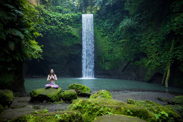 Young Caucasian woman meditating, practicing yoga at waterfall. Lotus pose. Hands in namaste mudra. Tibumana waterfall, Bali, Indonesia.
