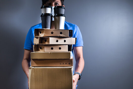 Courier In Facial Mask Holding Stack Of Boxes With Food And Beverage On Gray Background.