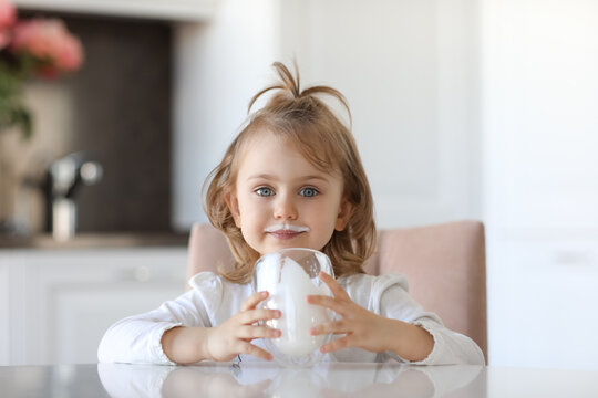 Blond cute baby girl with blue eyes with traces of milk on the lips is holding a glass of milk siting at a white table in a white kitchen. Milk for good health