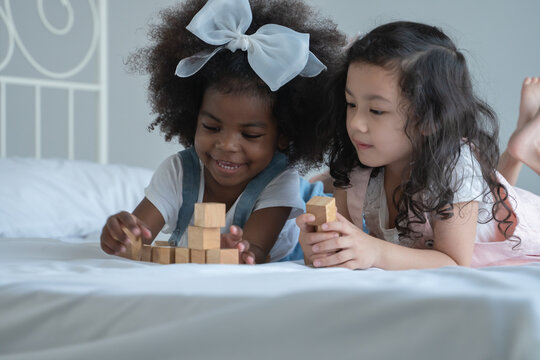 Two Little Girls, African And Asian Children Enjoy Playing Build Wooden Block Together On Bed At Bedroom 