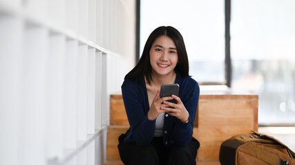 Cheerful young woman hands holding smart phone and smiling to camera while sitting in library.