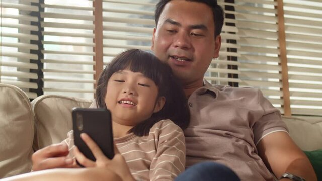 Happy Asian Family Dad and little kid daughter using a smart phone and having a video call chatting or taking a selfie sitting on a couch fun and enjoying online communication in living room at home.