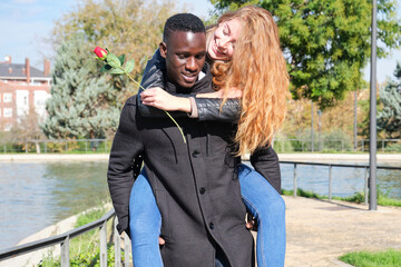 African black man and redhead caucasian woman enjoying piggyback ride with a rose. Young multiracial couple celebrating valentines day.