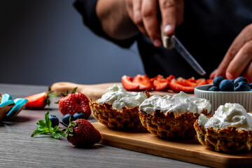 Hand with knife cuts strawberry. Berries on white cooking board. Fresh ingredients for a dessert. chef cooking berry dessert tarts with cream, strawberry and blueberry
