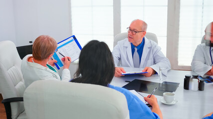 Obraz premium Mature woman doctor pointing on clipboard during briefing with coworkers working in hospital meeting room. Clinic expert therapist talking with colleagues about disease, medicine professional
