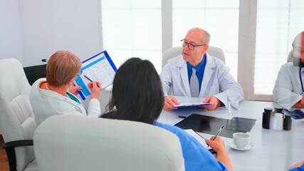 Mature woman doctor pointing on clipboard during briefing with coworkers working in hospital meeting room. Clinic expert therapist talking with colleagues about disease, medicine professional